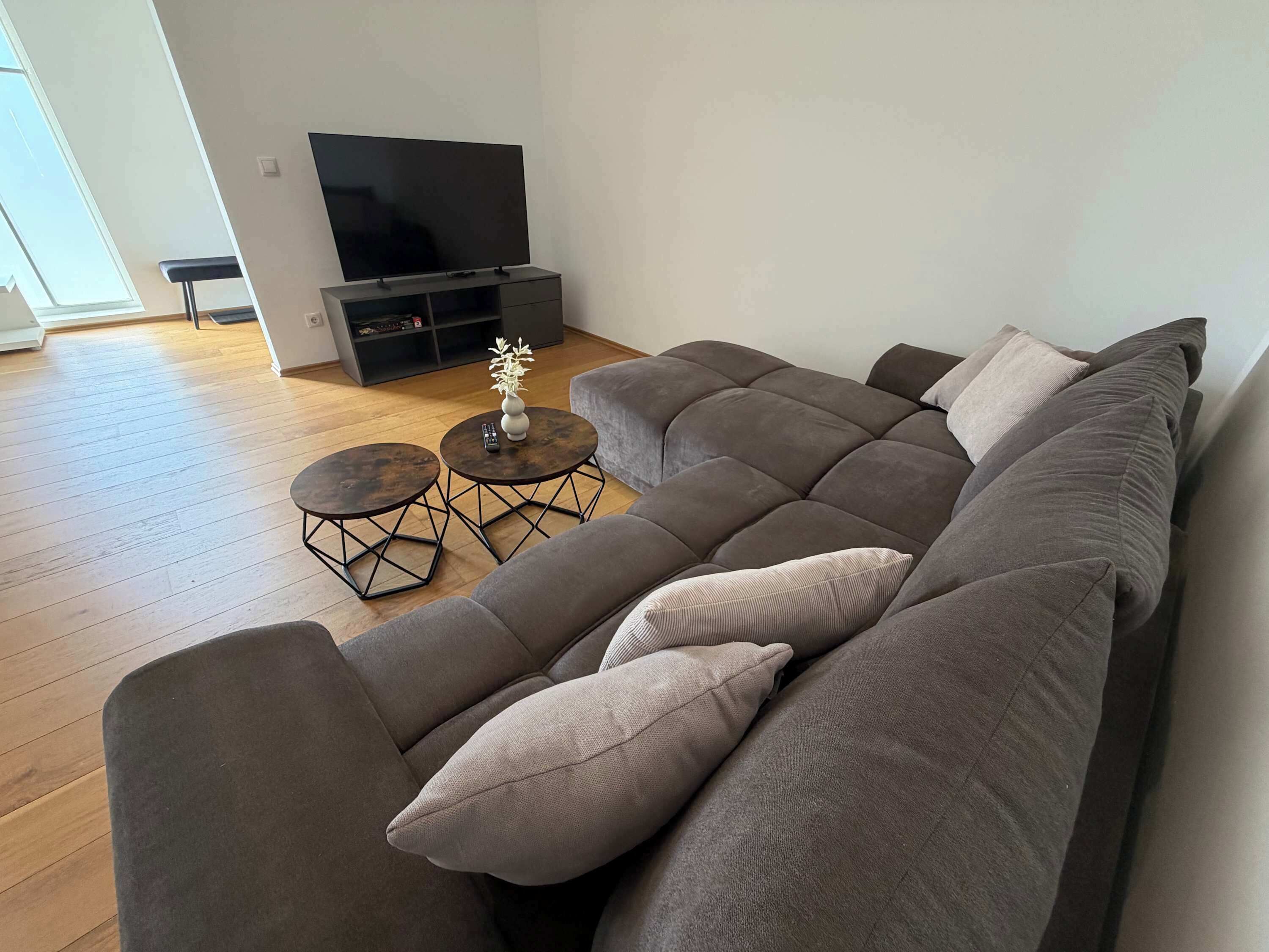Un salon moderne avec un canapé d’angle, deux petites tables rondes et un téléviseur à écran plat posé sur un meuble.