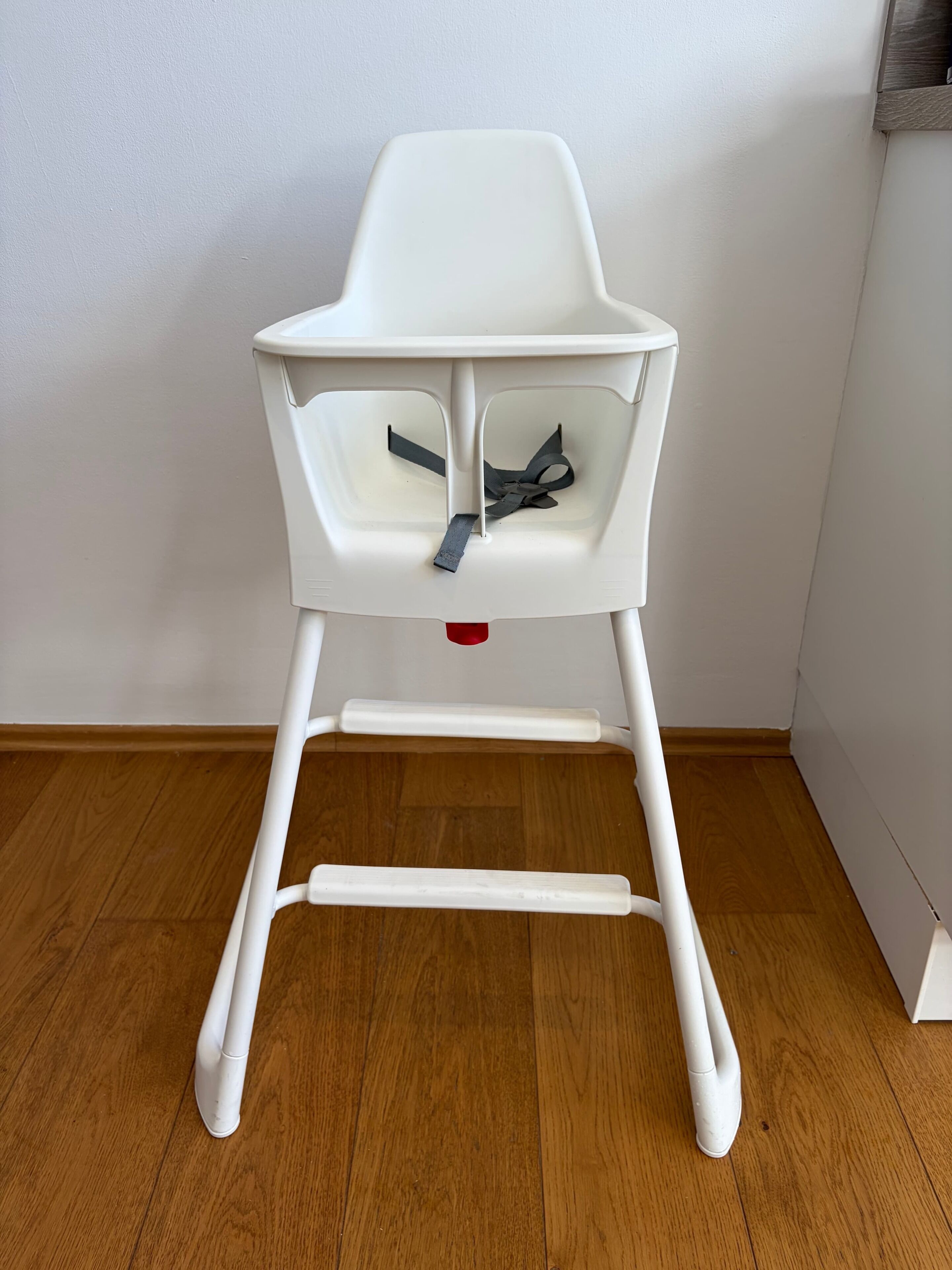 Una silla alta blanca con bandeja, sobre un piso de madera frente a una pared blanca.