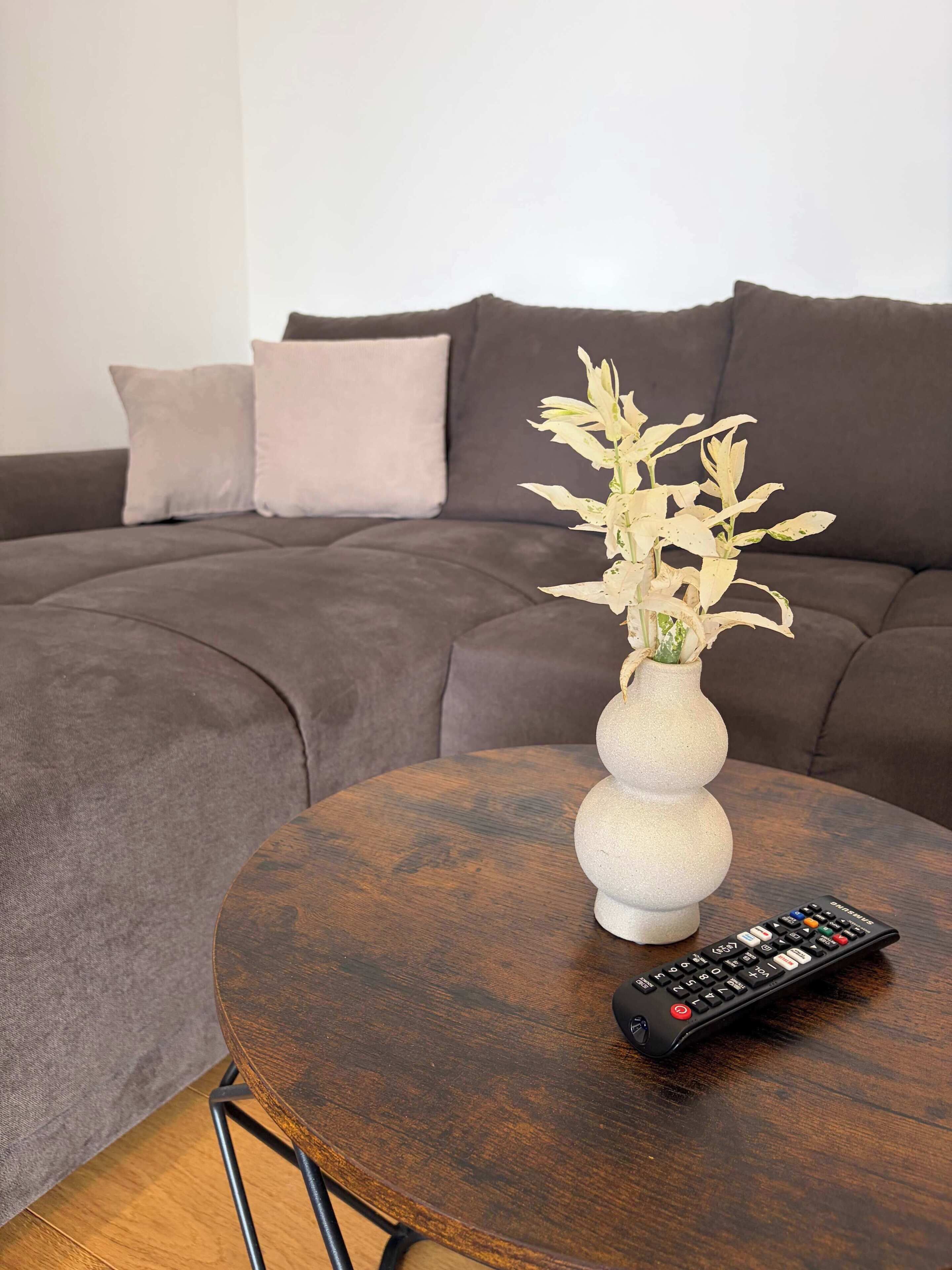 A round wooden table with a vase of white flowers and a remote control.