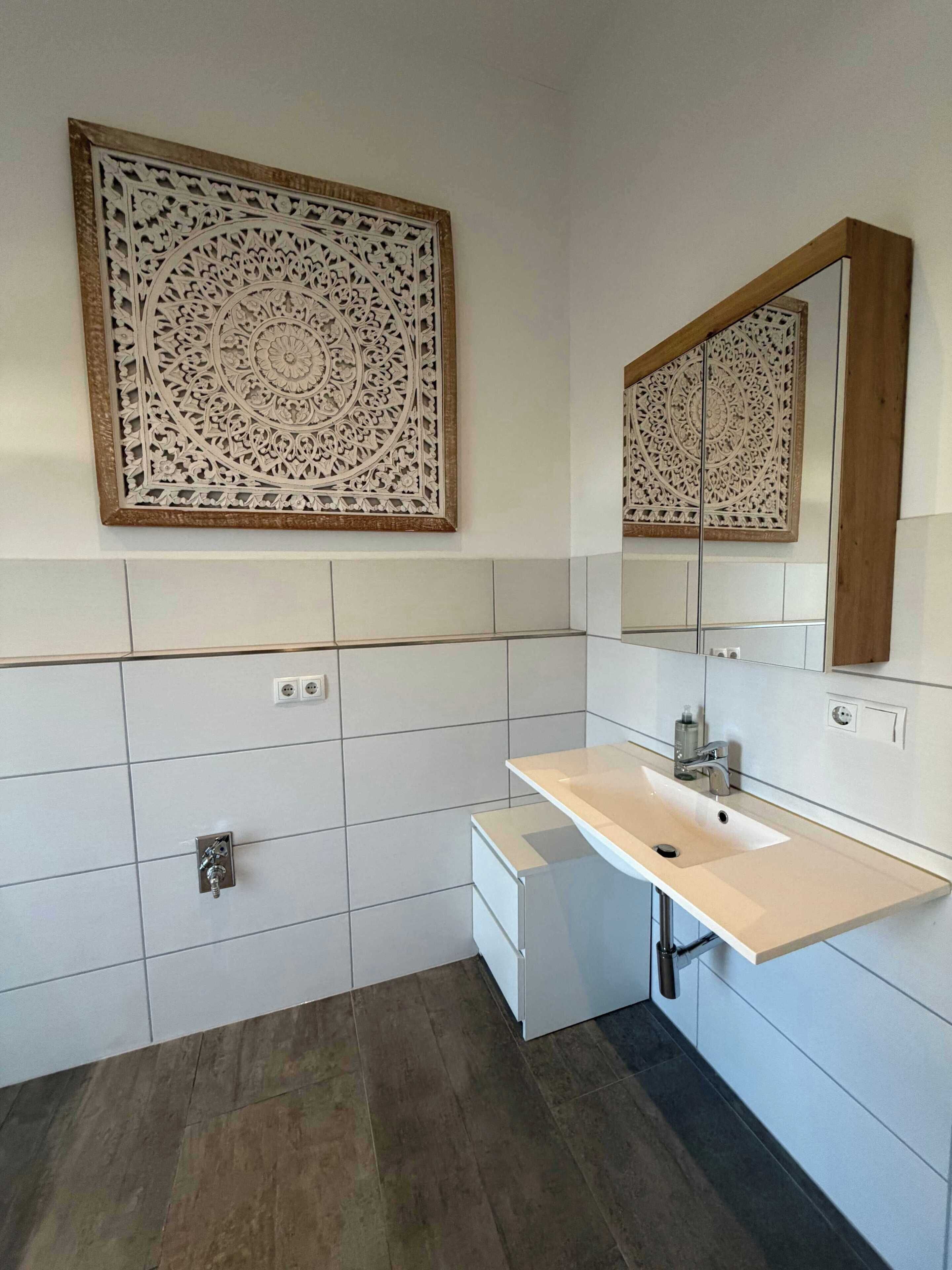 Ein Badezimmer mit weiß gefliester Wand, einem beigen Waschbecken, einem Spiegel mit Holzrahmen und einer dekorativen Wandverkleidung.