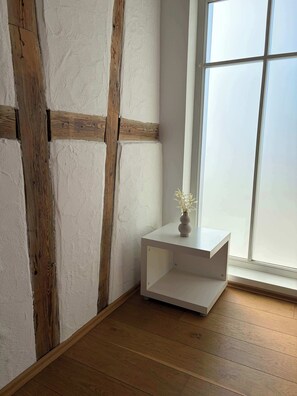 A room with a wooden floor, a white wall with exposed beams, a small white side table with a vase, and a large window.