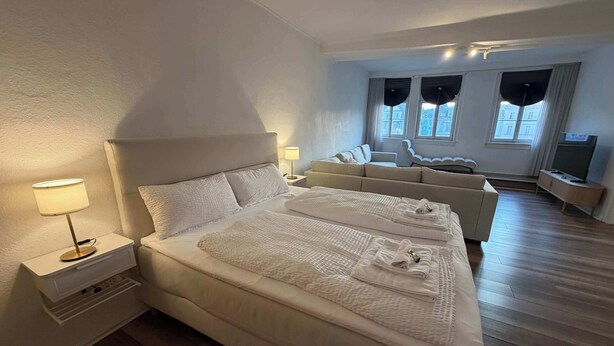Quarto de hotel moderno com uma cama grande, uma mesinha de cabeceira com luminária, um sofá, uma TV e vista para a cidade pelas janelas.