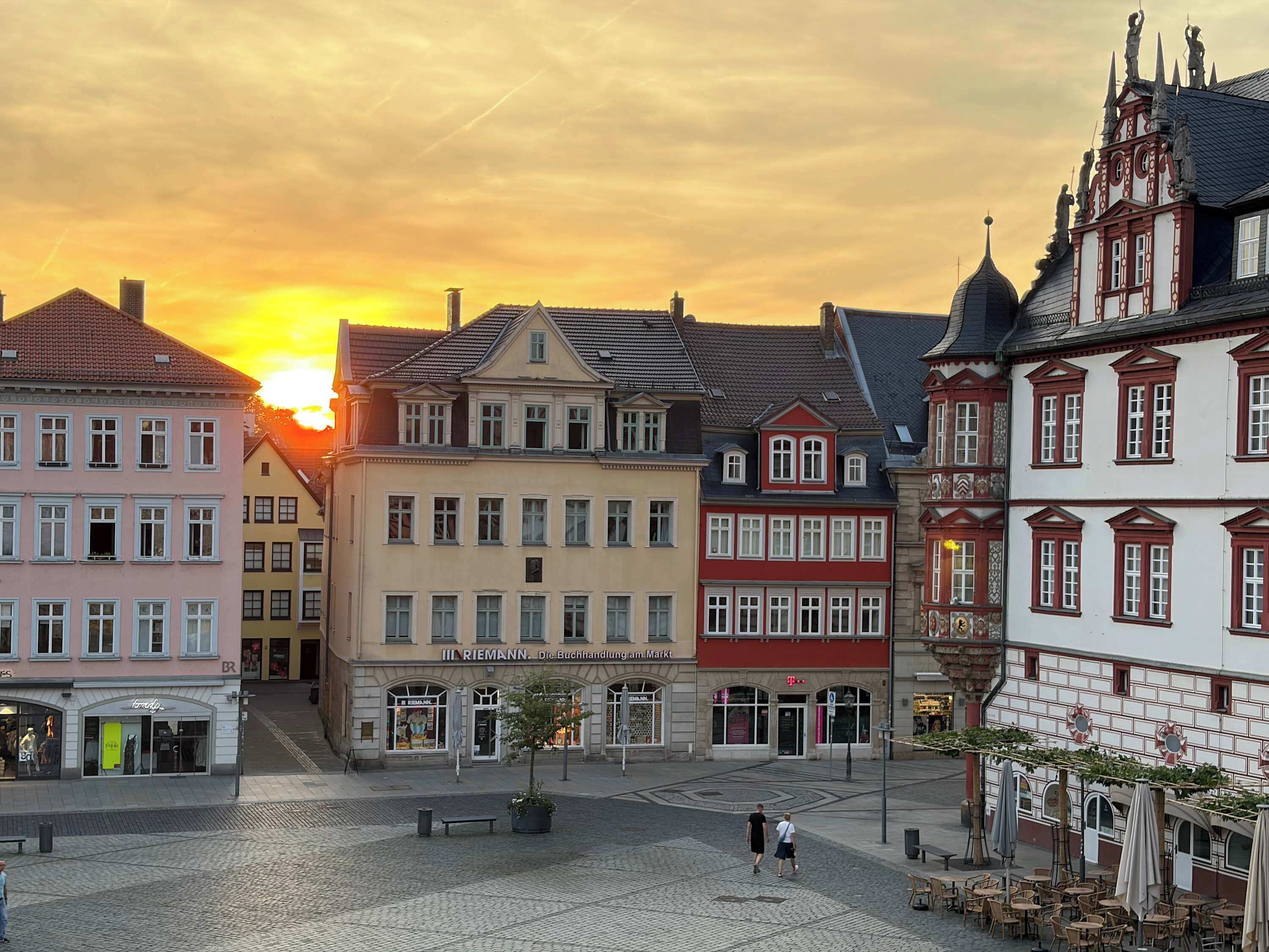 Een Europees stadsplein met historische gebouwen, een open ruimte in het midden en een ondergaande zon op de achtergrond.
