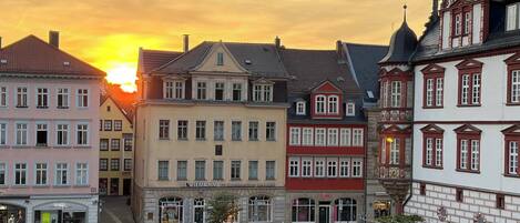 Een Europees stadsplein met historische gebouwen, een open ruimte in het midden en een ondergaande zon op de achtergrond.