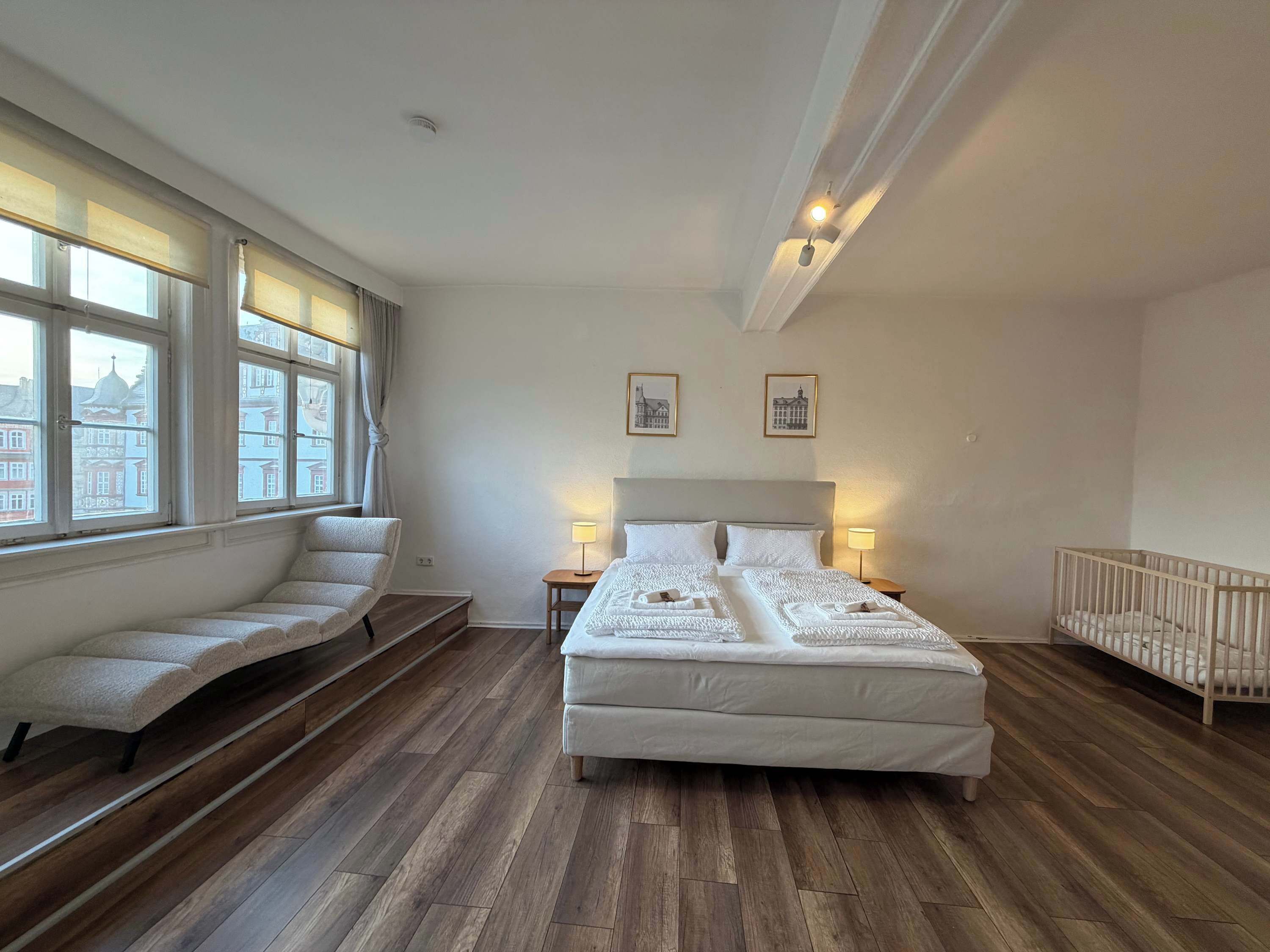 Une chambre d’hôtel avec un grand lit, un coin salon avec un banc, un berceau et une vue sur les bâtiments par les fenêtres.
