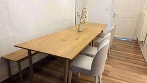 Uma área de refeições com uma mesa de madeira, quatro cadeiras cinzentas e um vaso com flores.