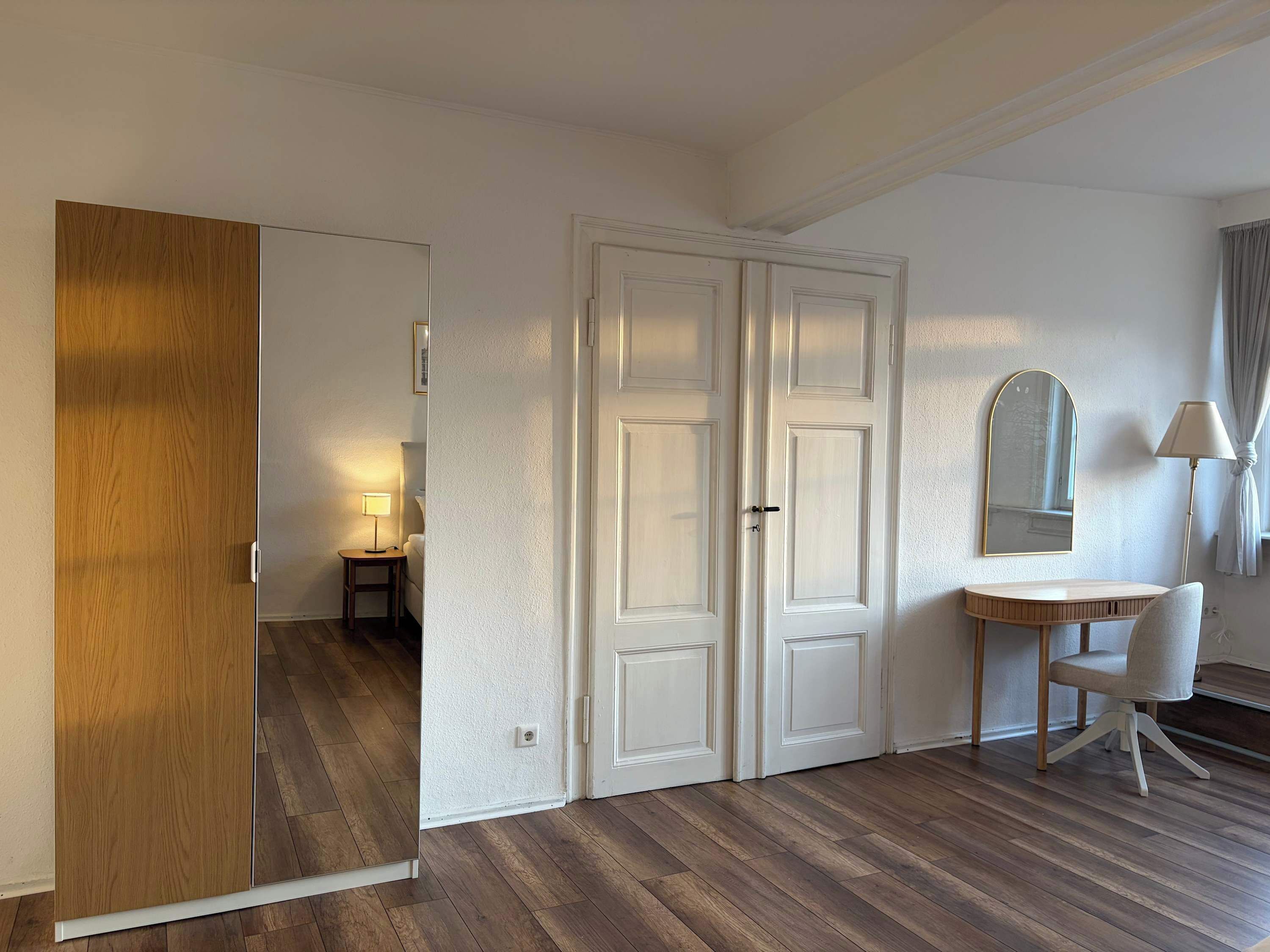 A room with a wooden wardrobe, a white door, a small bedside table with a lamp, a mirror, and a chair.