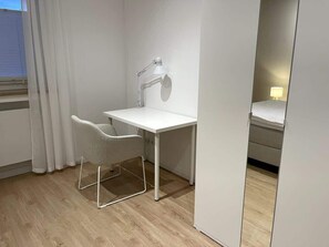 Ein kleiner, moderner Raum mit weißem Schreibtisch, einem Stuhl, einer Schreibtischlampe und einem Spiegel.