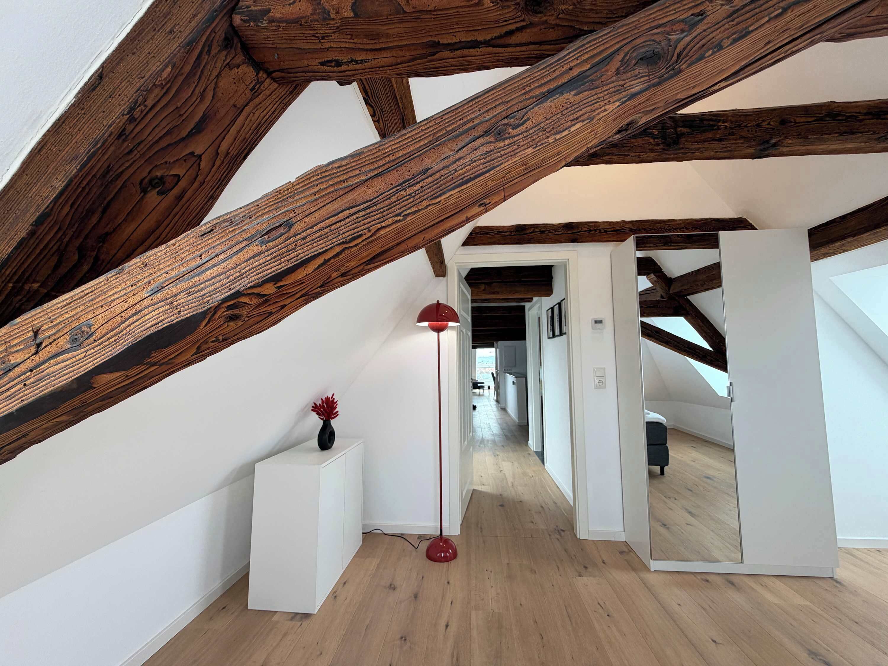 Une pièce avec des poutres apparentes, une armoire blanche, un lampadaire rouge et un couloir menant à une autre pièce.