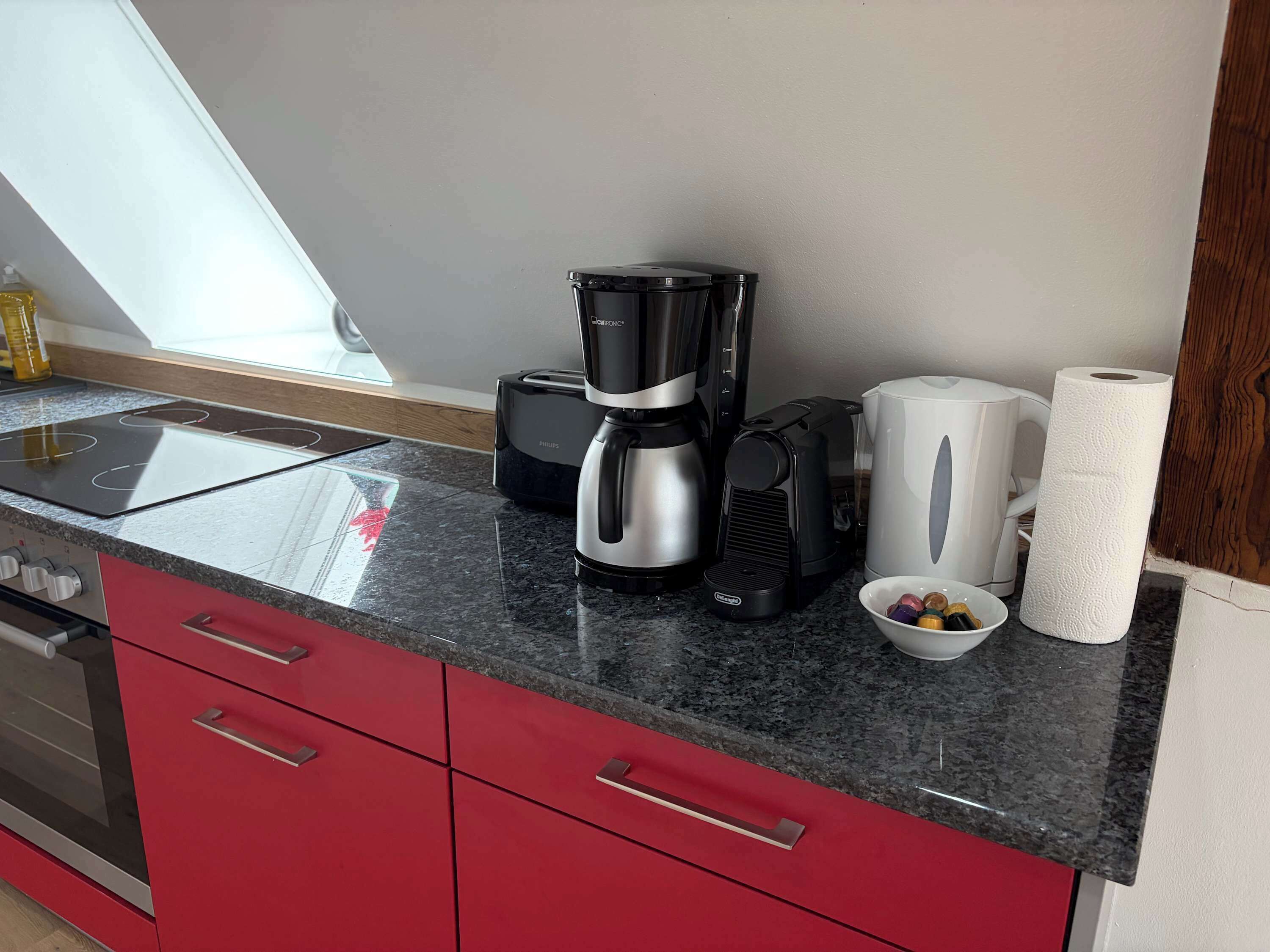 Une cuisine au design moderne, avec une machine à café noire et argentée, une bouilloire blanche et un saladier rempli de fruits posé sur le plan de travail.