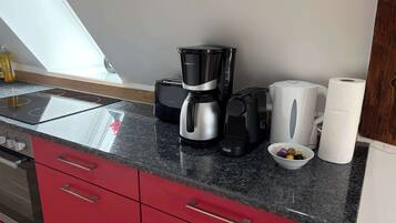 Une cuisine au design moderne, avec une machine à café noire et argentée, une bouilloire blanche et un saladier rempli de fruits posé sur le plan de travail.