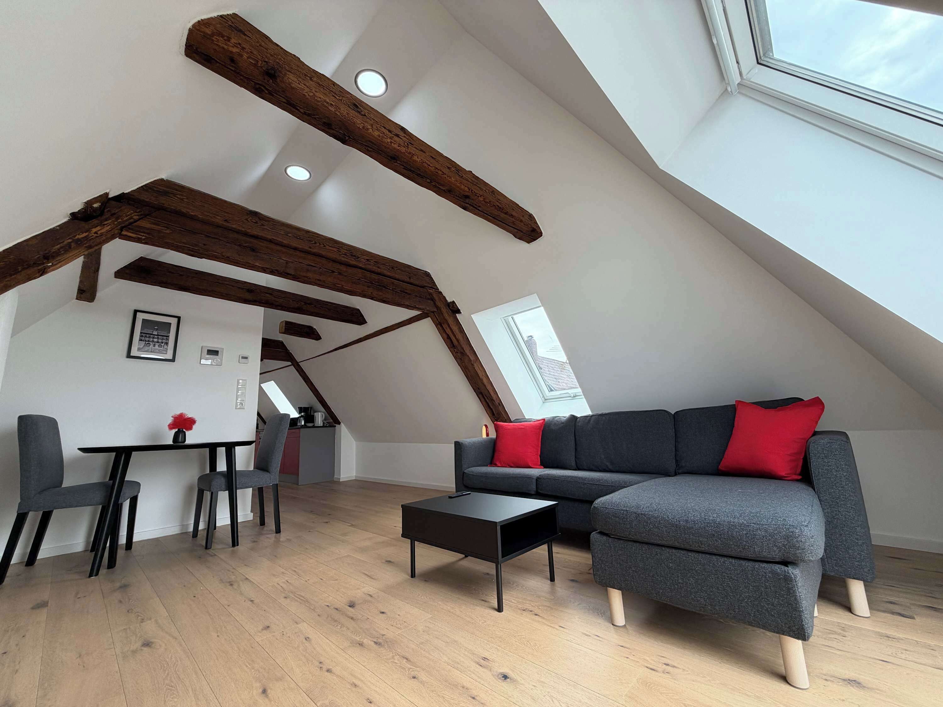 A loft-style living space with exposed wooden beams, a gray sectional sofa with red cushions, a small black coffee table, and a dining area with chairs and a table.