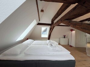 Ein Zimmer mit schräg verlaufender Decke, Holzbalken, einem Bett mit weißen Leinen, einem kleinen Tisch mit einer Vase und einer roten Stehlampe.