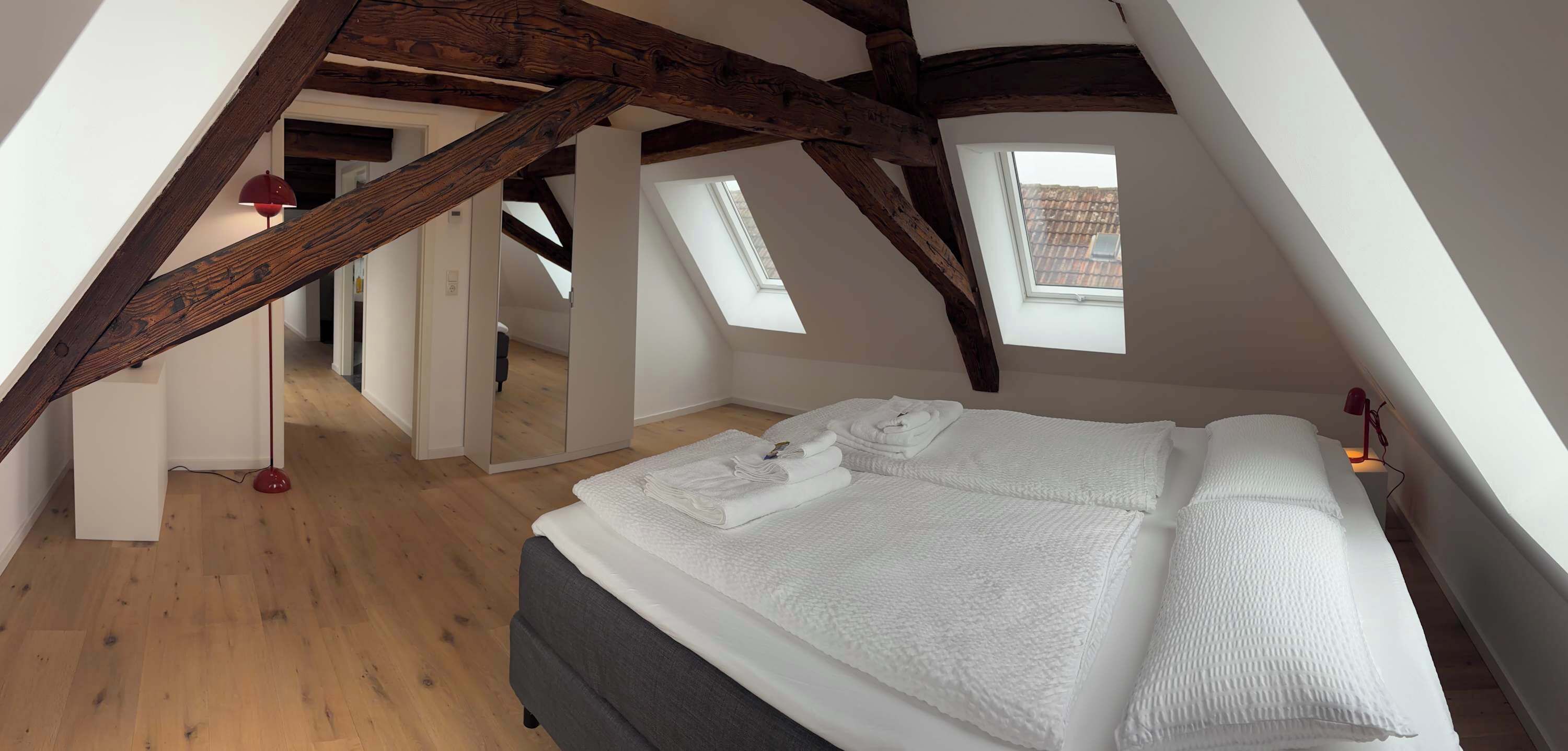 Un dormitorio con techo inclinado, vigas de madera, una cama con ropa de cama blanca y un tragaluz.