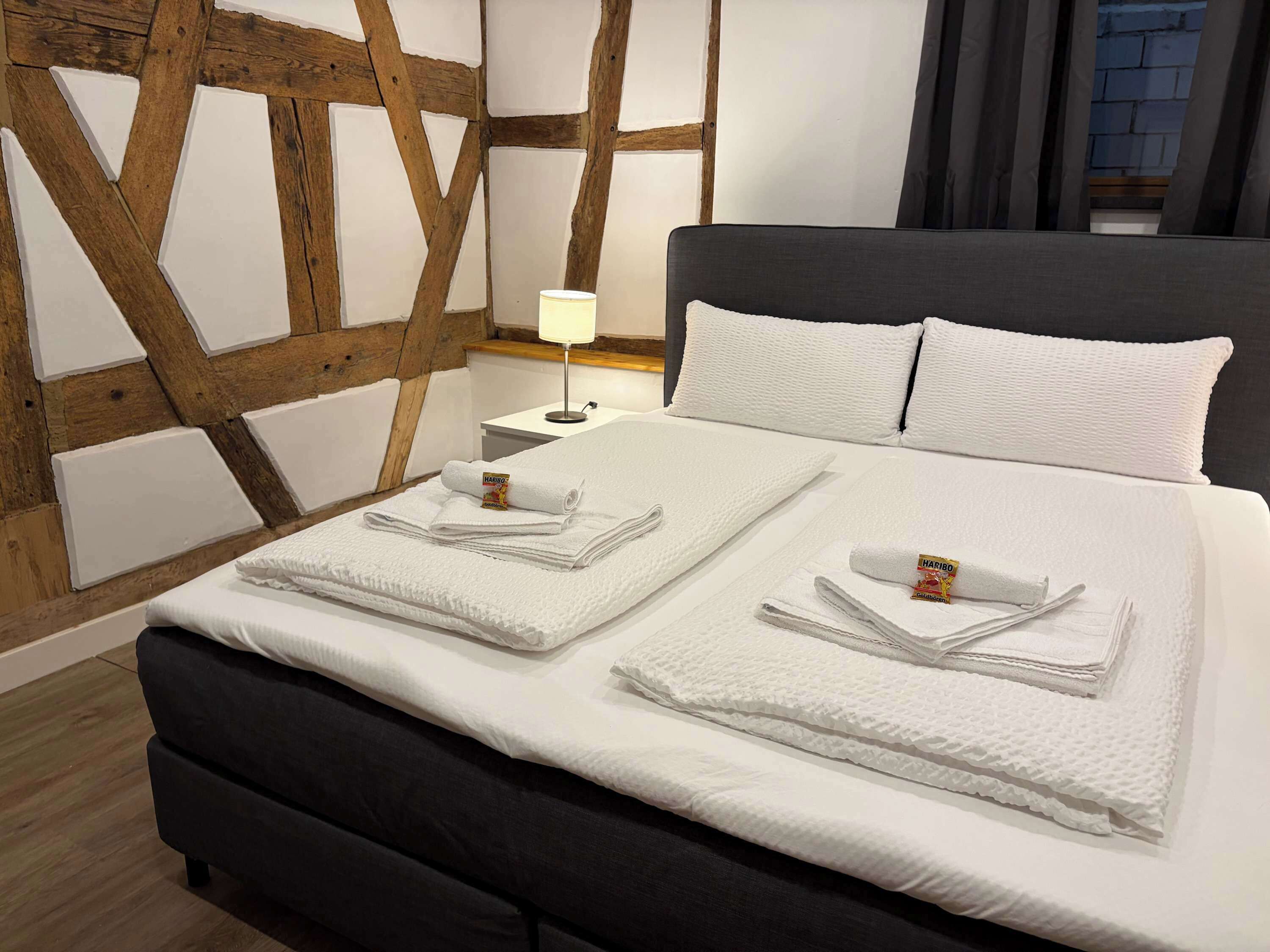 Een netjes opgemaakt bed met witte lakens en twee gevouwen handdoeken met Haribo logo, een bedlamp en een houten wand met zichtbare balken.