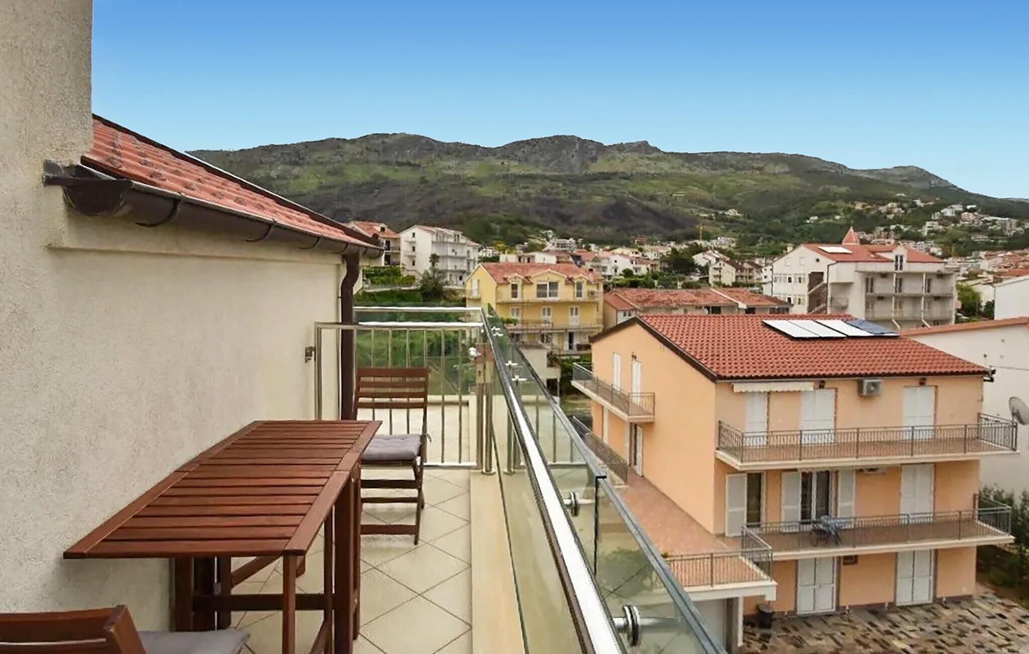 Un balcón con mesa y sillas, con vista a una zona residencial con casas y una montaña al fondo.
