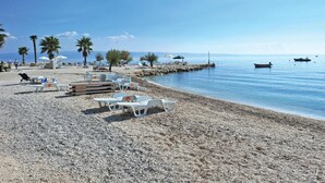 Een kiezelstrand met ligbedden en parasols, een rotsachtige kustlijn, palmbomen en boten op het water.