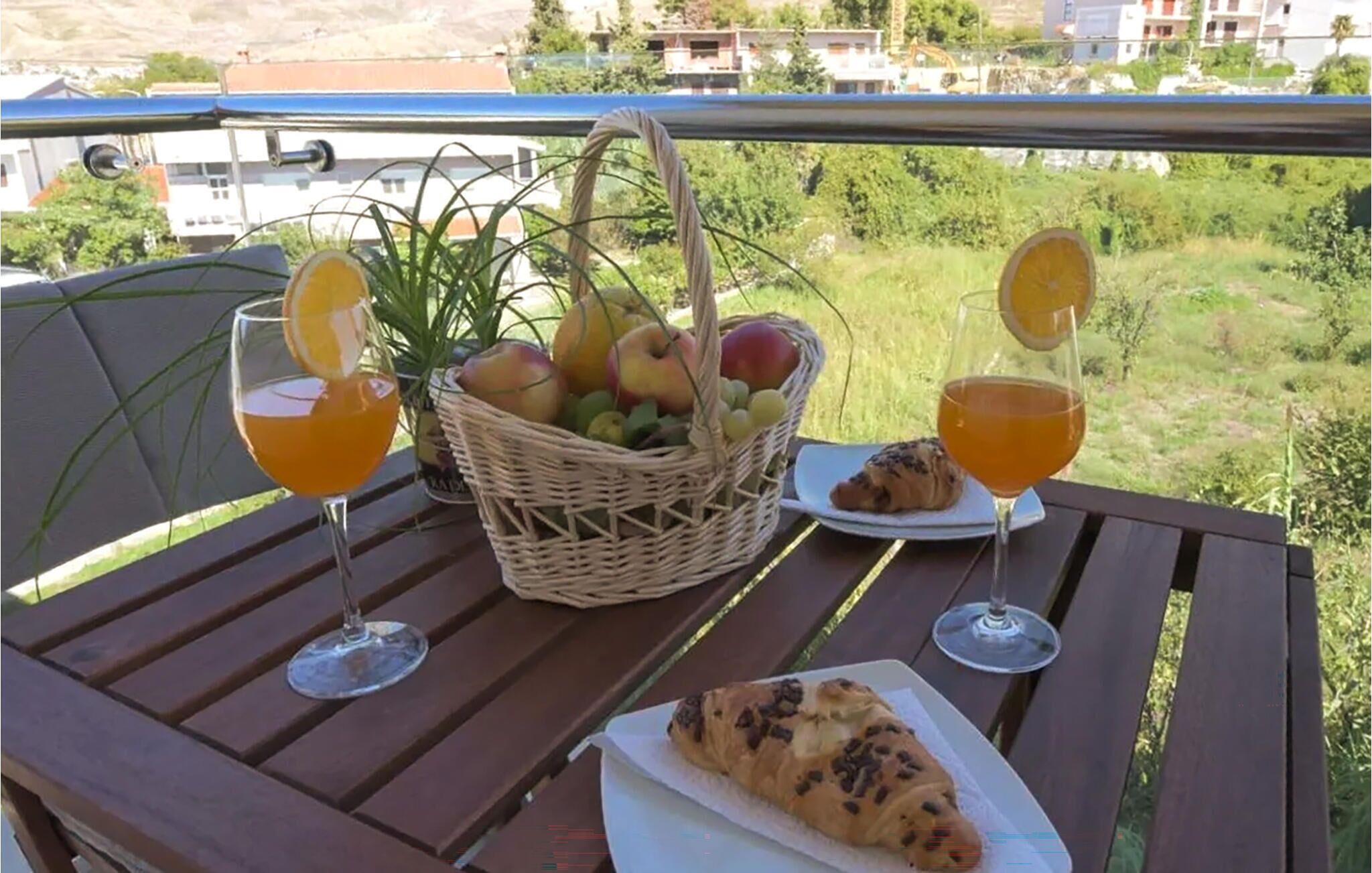 A table with a basket of fruit, two glasses of orange juice, a croissant, and a plate of food on a balcony overlooking a green landscape.