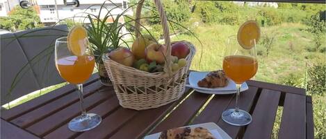 A table with a basket of fruit, two glasses of orange juice, a croissant, and a plate of food on a balcony overlooking a green landscape.