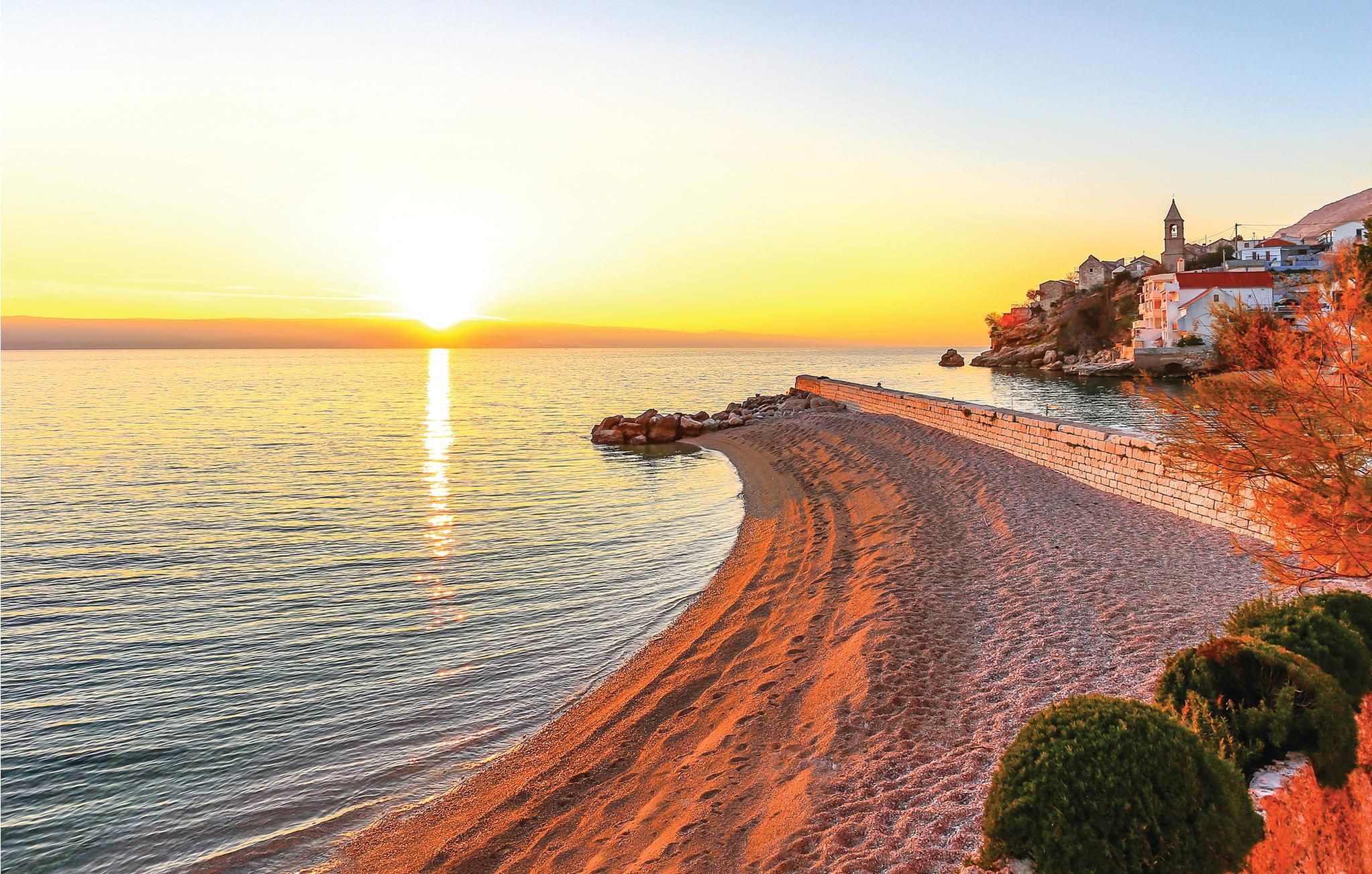 A coastal sunset view with a stone wall leading to a beach, a church on a hill, and buildings along the shore.