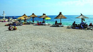 Ein Strand mit Sonnenschirmen und Liegestühlen, eine felsige Küstenlinie und ein Kruzifix im Hintergrund.
