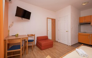 Una habitación compacta con una cama, una mesita con sillas, un televisor en la pared, una kitchenette y una puerta.