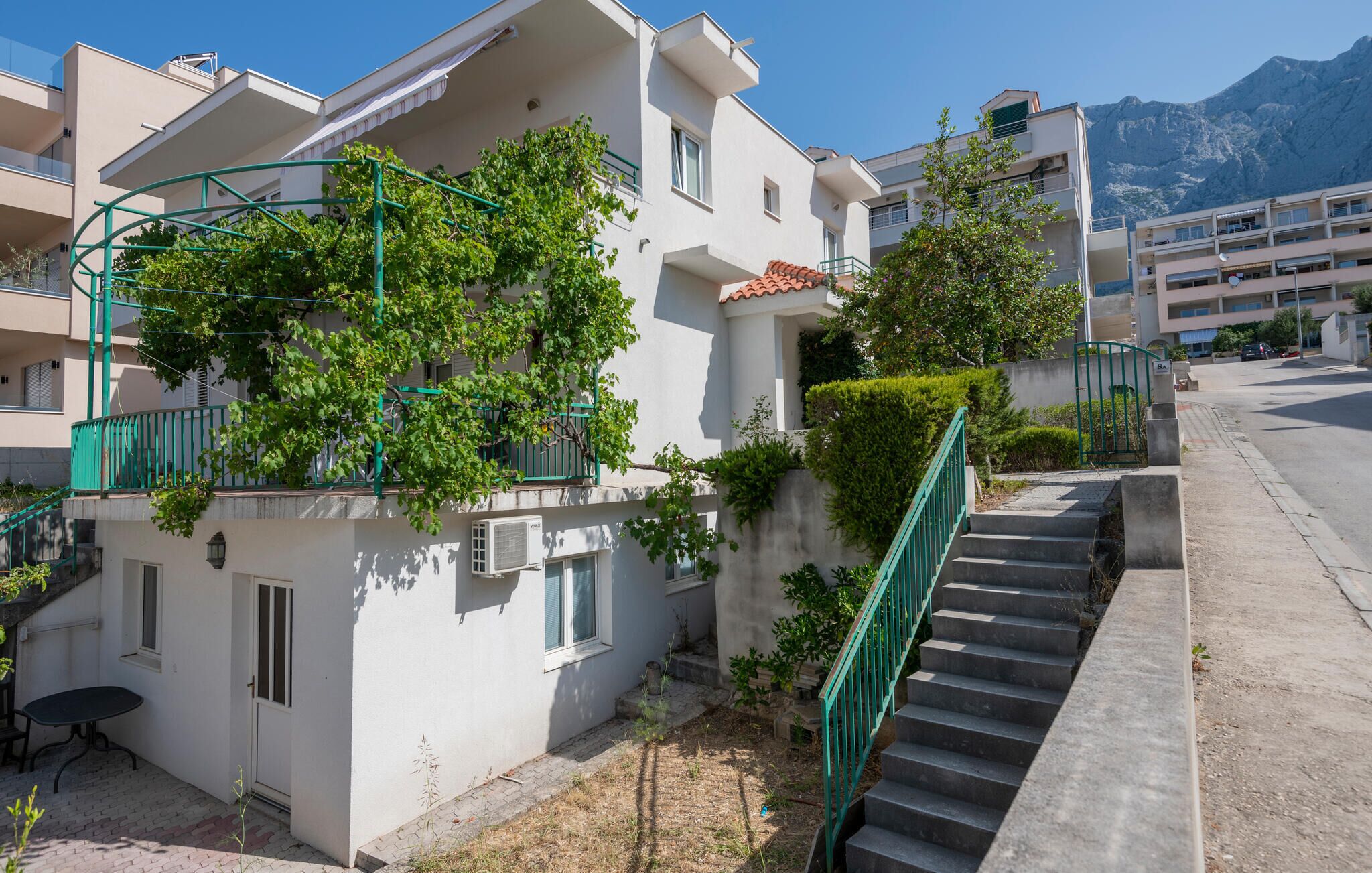 A residential building with a staircase leading to an upper floor, surrounded by greenery and a mountain in the background.