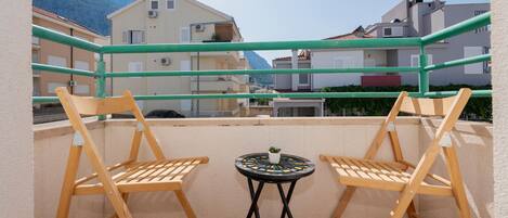 A balcony with two wooden chairs and a small table, overlooking a residential area with buildings and a mountain in the background.