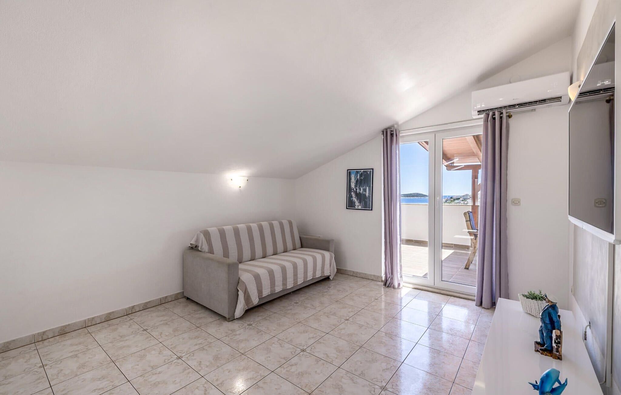 Une pièce comprenant un canapé, un sol carrelé, une porte-fenêtre coulissante donnant sur un balcon et un climatiseur fixé au mur.