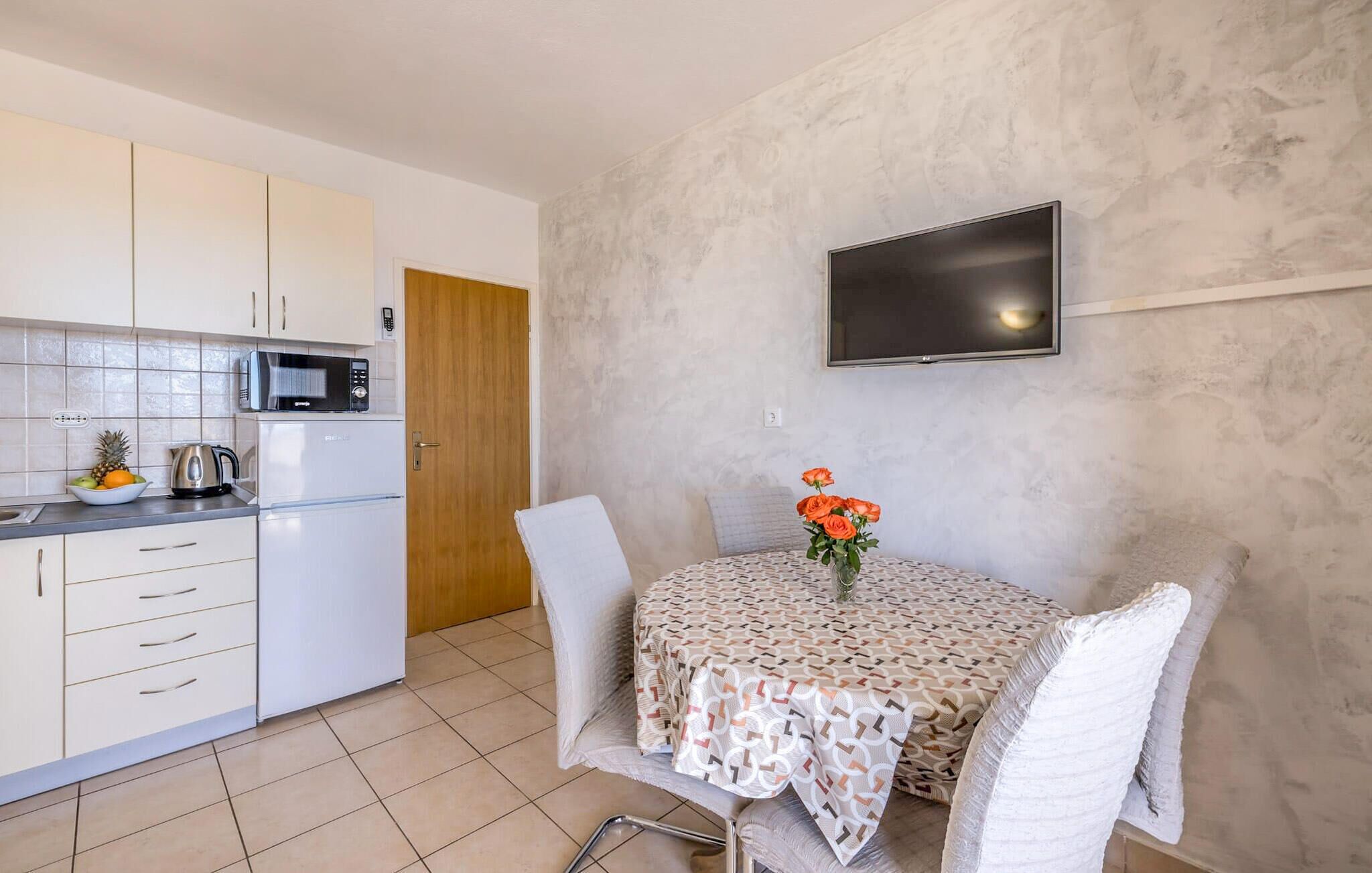 A compact kitchen with white cabinets, a microwave, and a kettle on the counter. There's a dining table with a patterned tablecloth and chairs. A flat-screen TV is mounted on the wall above the table.
