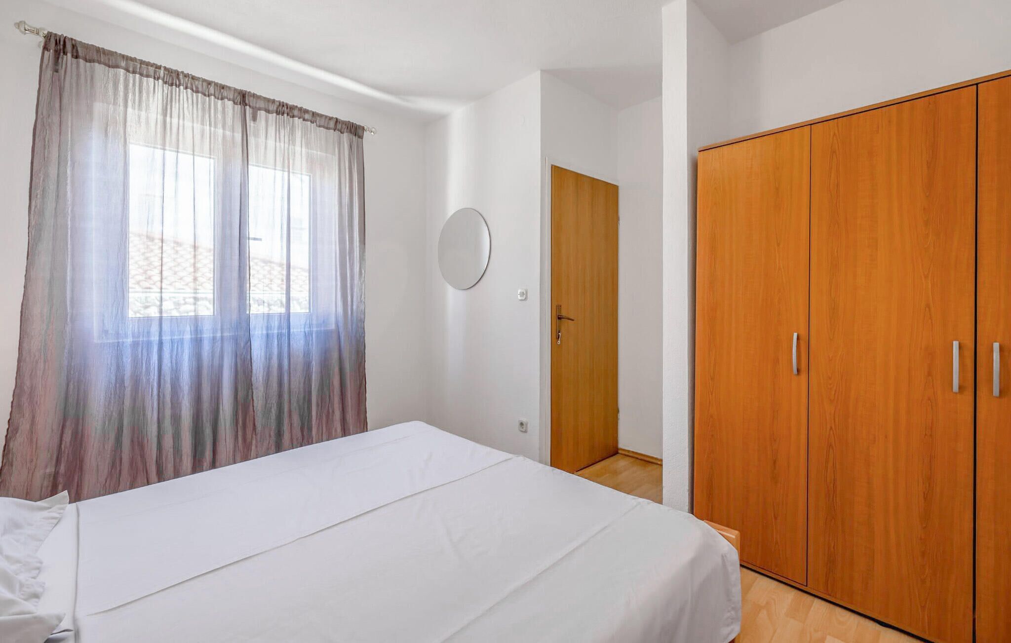 A bedroom with a bed, a wooden wardrobe, a window with sheer curtains, and a door.