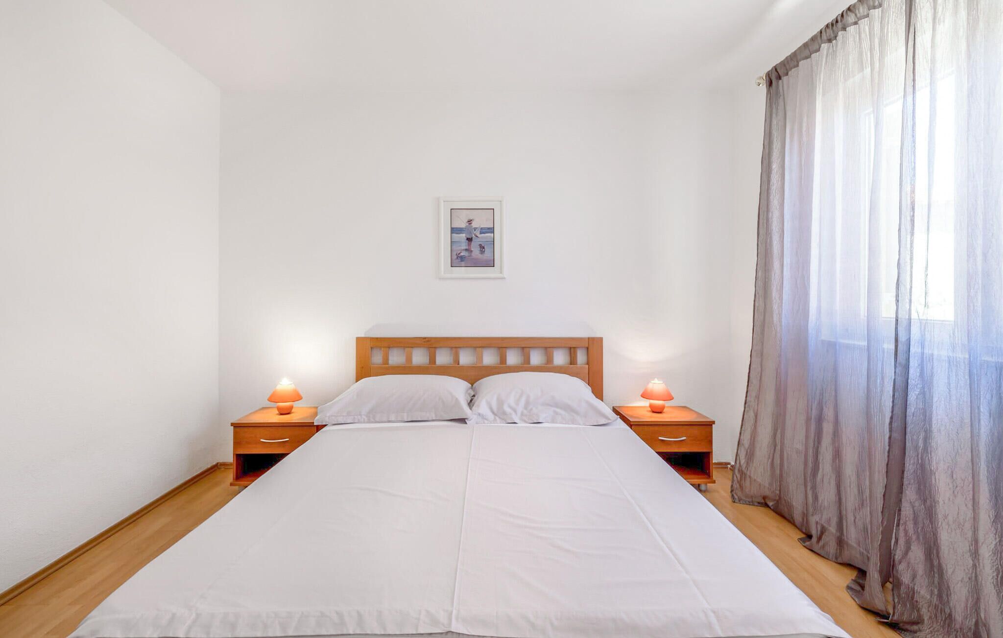 Ein Einzelbett mit hölzernem Kopfteil, zwei Nachttische mit Lampen, ein gerahmtes Bild an der Wand und ein Fenster mit durchsichtigen Vorhängen.