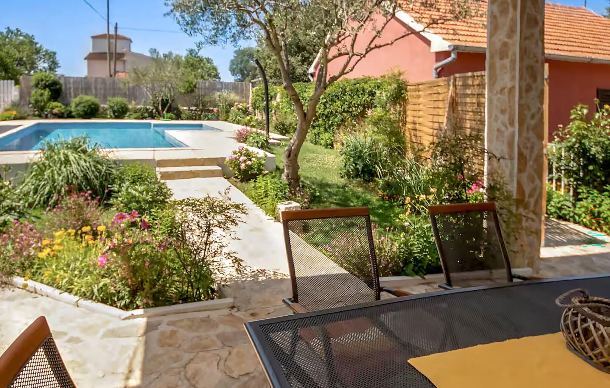 Un espace piscine agrémenté d’une table et de chaises sur une terrasse, le tout entouré d’un jardin où poussent différentes plantes et fleurs.