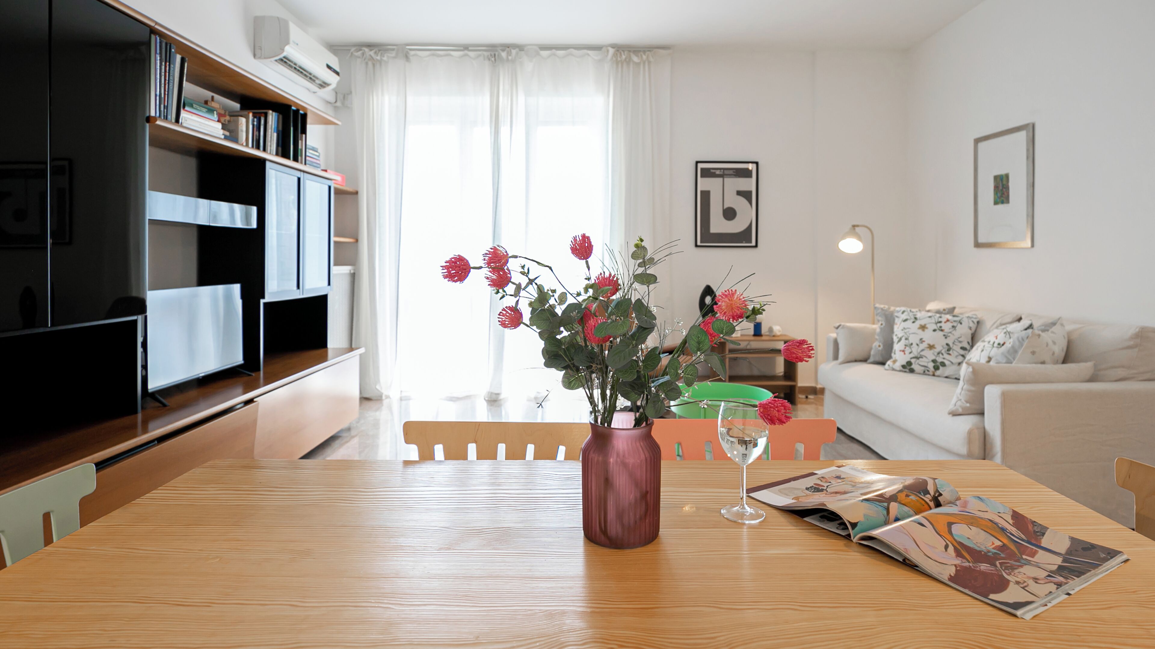 A modern living room with a wooden dining table, a vase with flowers, and a magazine.
