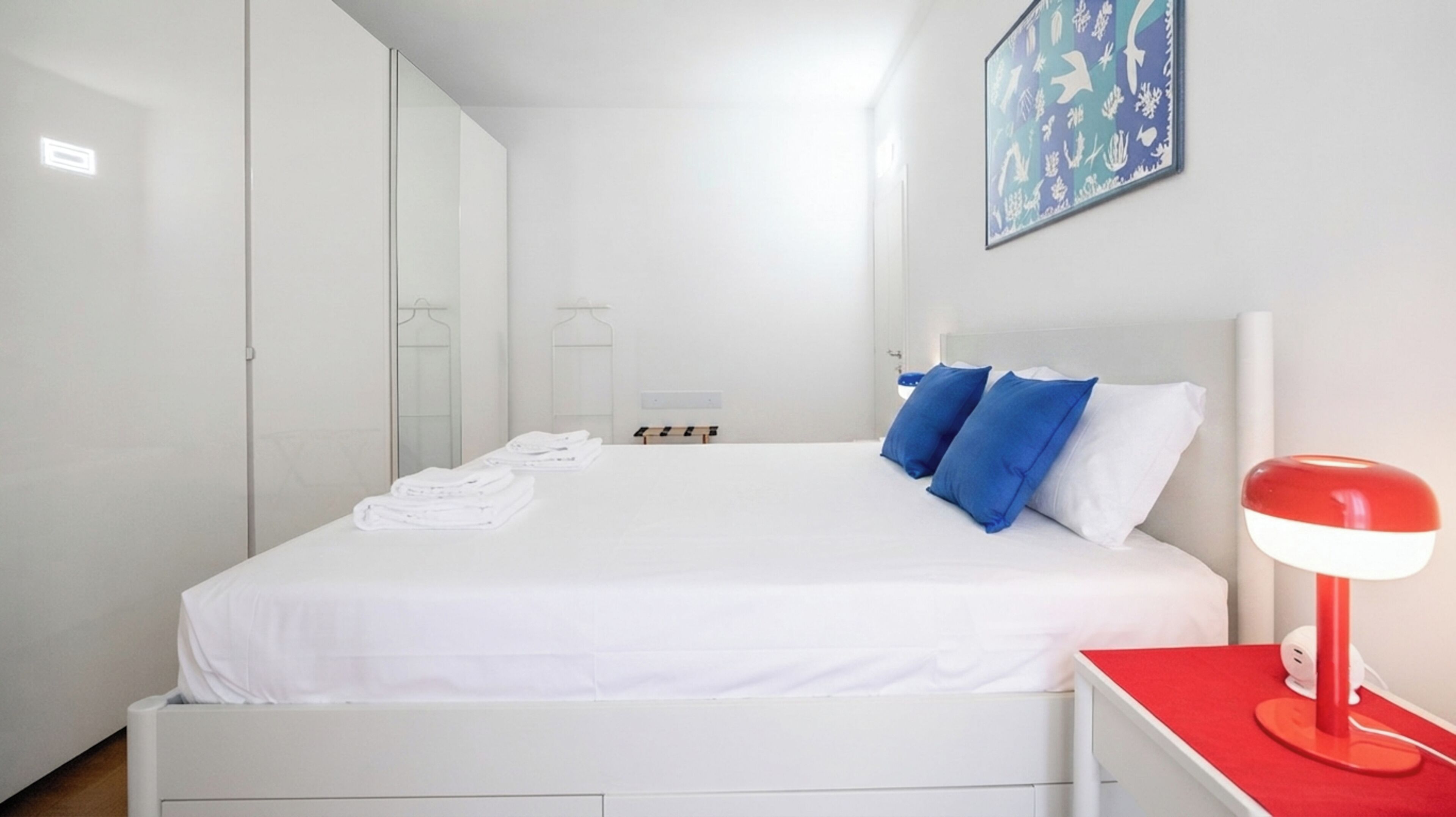 Habitación de hotel con cama, ropa blanca de cama, lámpara de noche roja y una obra de arte colgada en la pared.