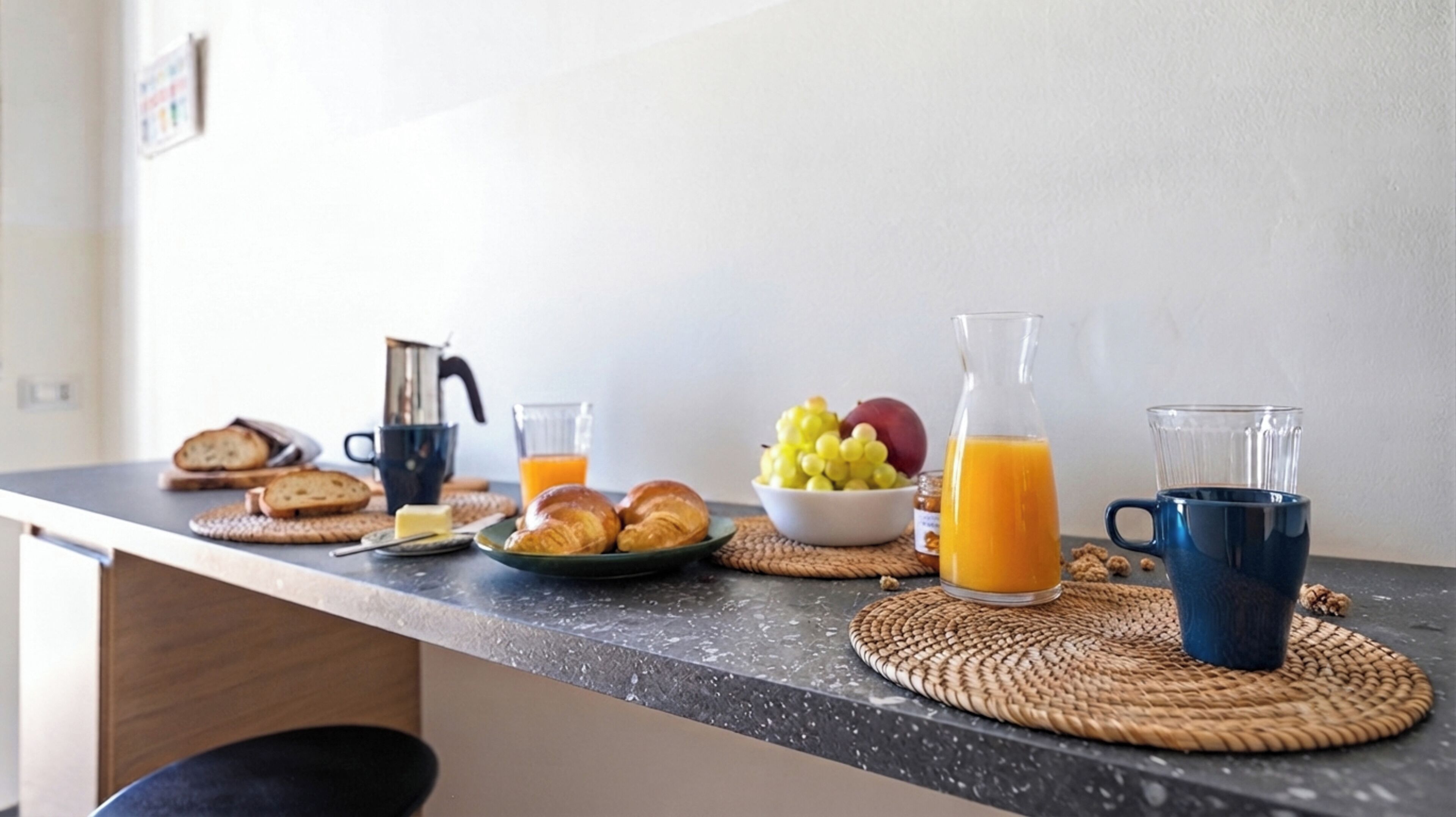 Una mesa de desayuno con cruasanes, un bol de fruta, una jarra de jugo de naranja y una cafetera.
