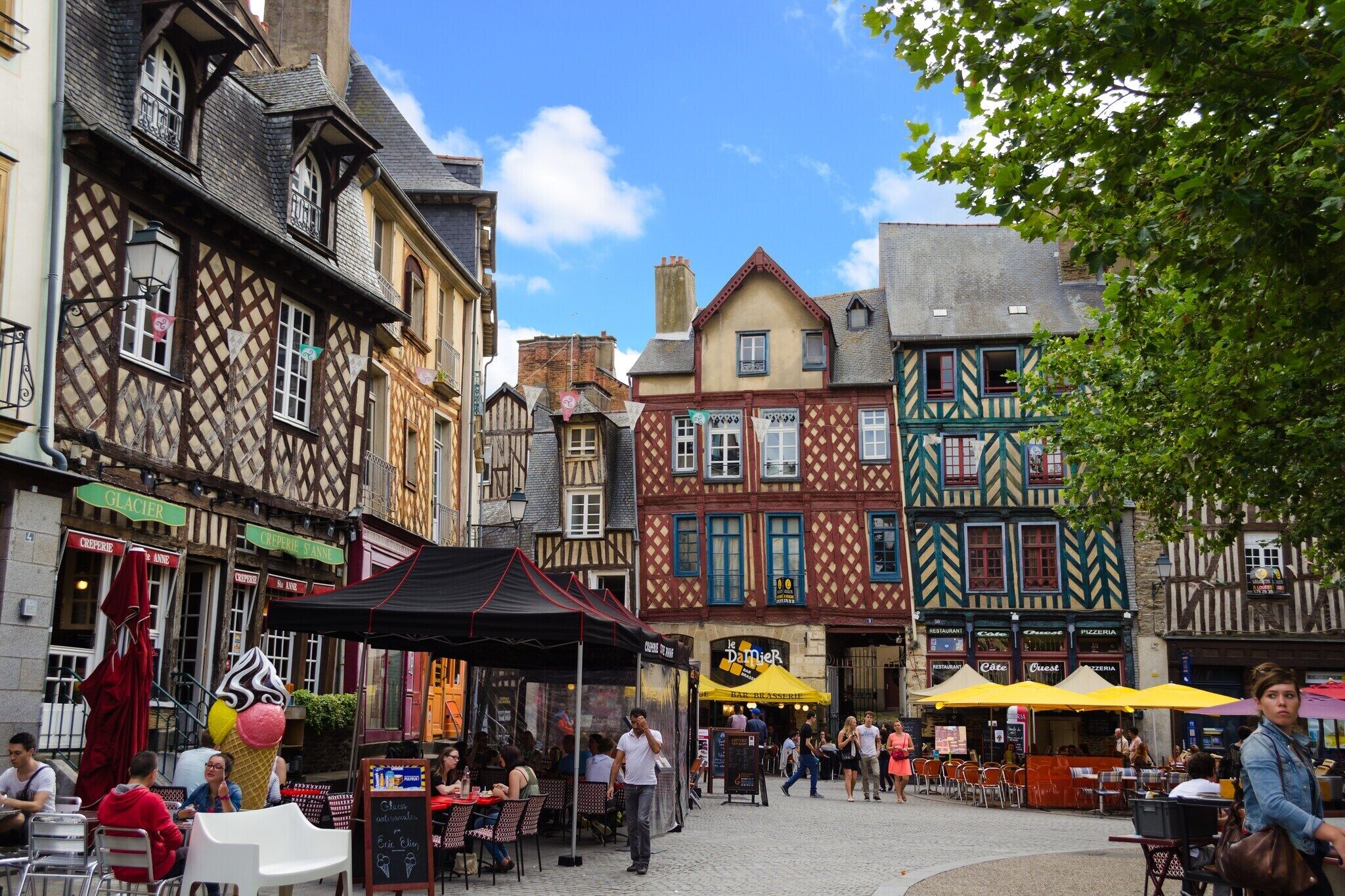 A cobblestone street with traditional half-timbered buildings, outdoor seating at cafes, and a clear blue sky.