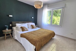 Ein Schlafzimmer mit Bett, Nachttisch, Lampe, Fenster und Blick auf Grünflächen.