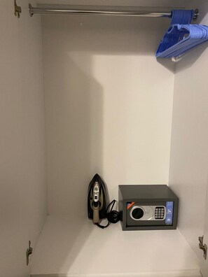 Um quarto pequeno e vazio, com teto de barra de metal, ferro de passar preso na parede e um pequeno aparelho eletrônico em uma prateleira.
