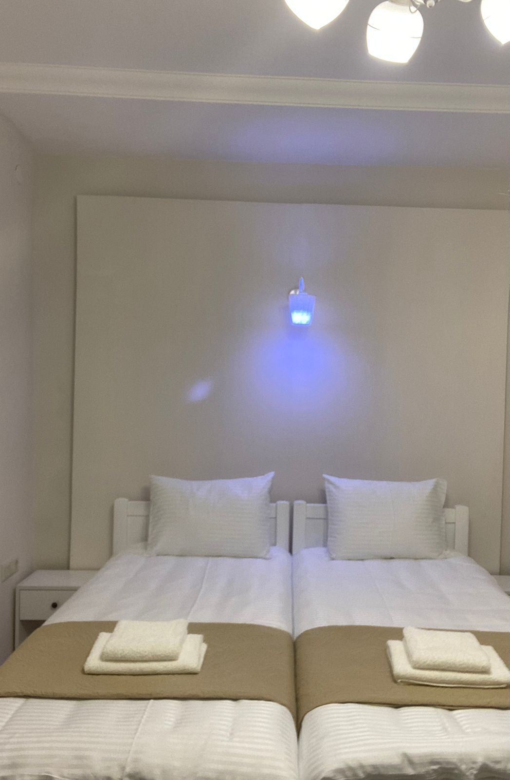 A hotel room with two beds, each with a white headboard, a brown bedspread, and a white pillow. There is a small blue light fixture on the ceiling.