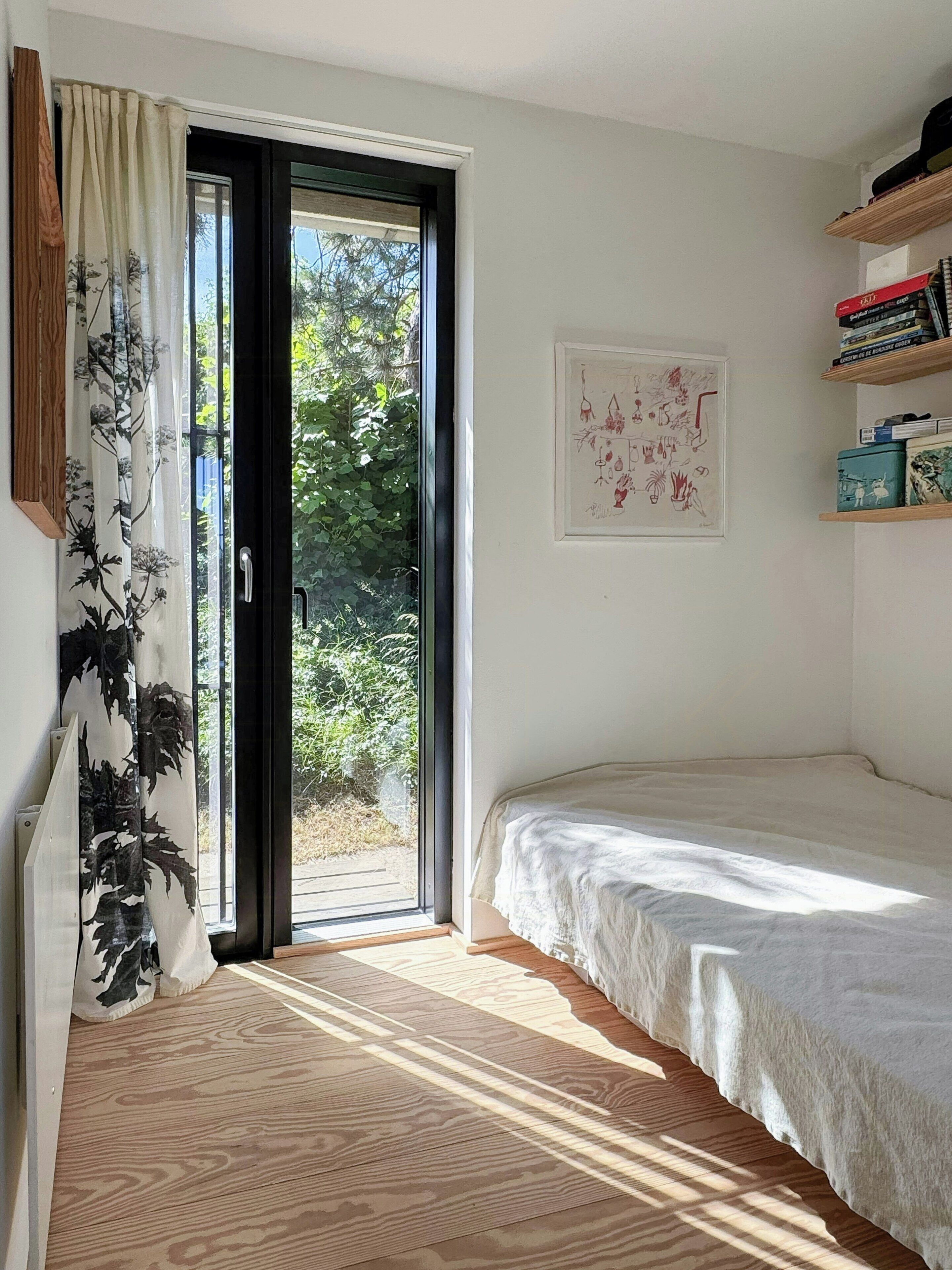 Ein Schlafzimmer mit einem Bett, einer Tür zum Balkon, einem gerahmten Kunstwerk an der Wand und einem Regal mit Büchern.