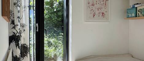 A bedroom with a bed, a door leading to a balcony, a framed artwork on the wall, and a shelf with books.