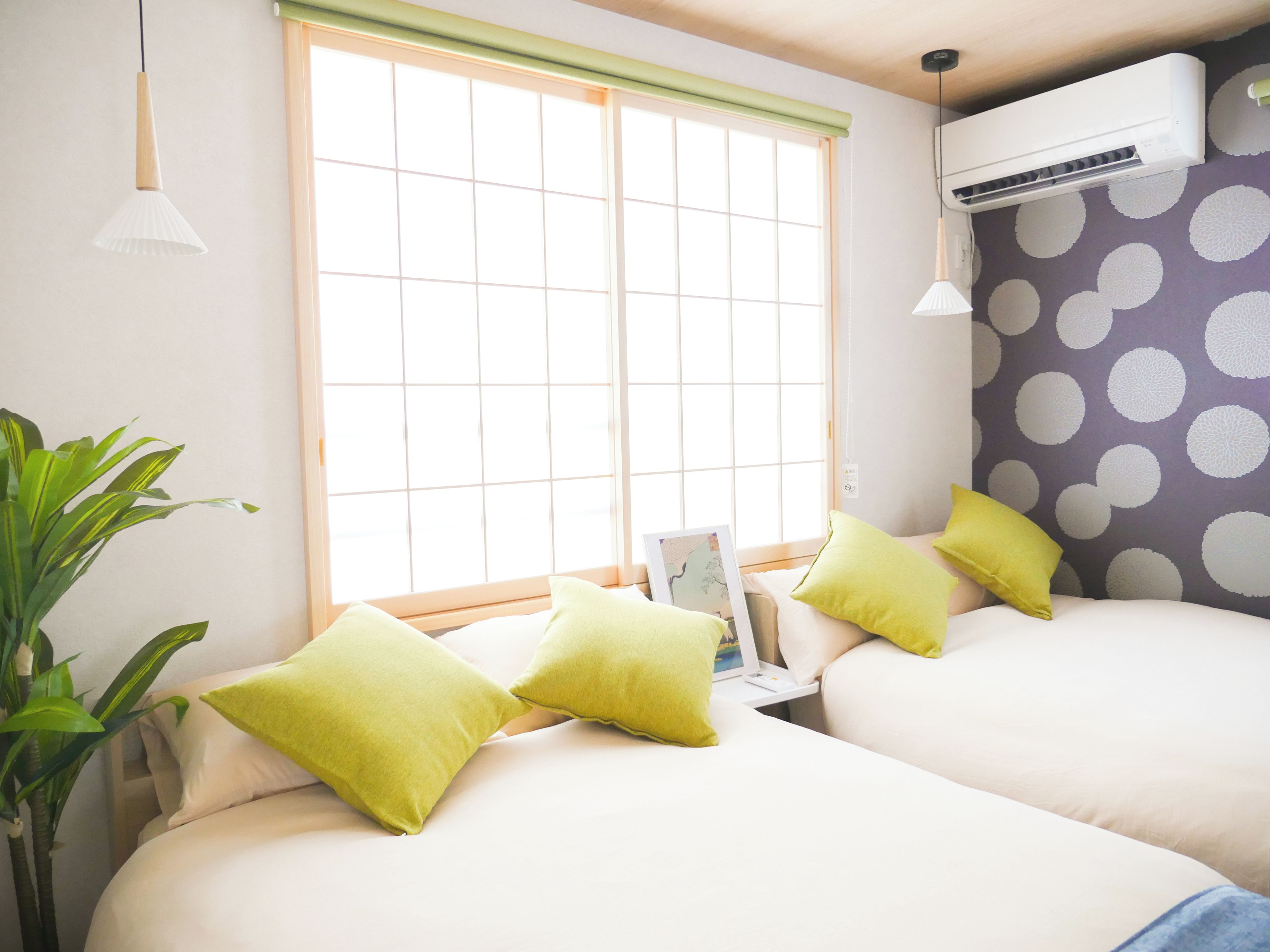 A room with two beds, a window with blinds, an air conditioner, and a potted plant.
