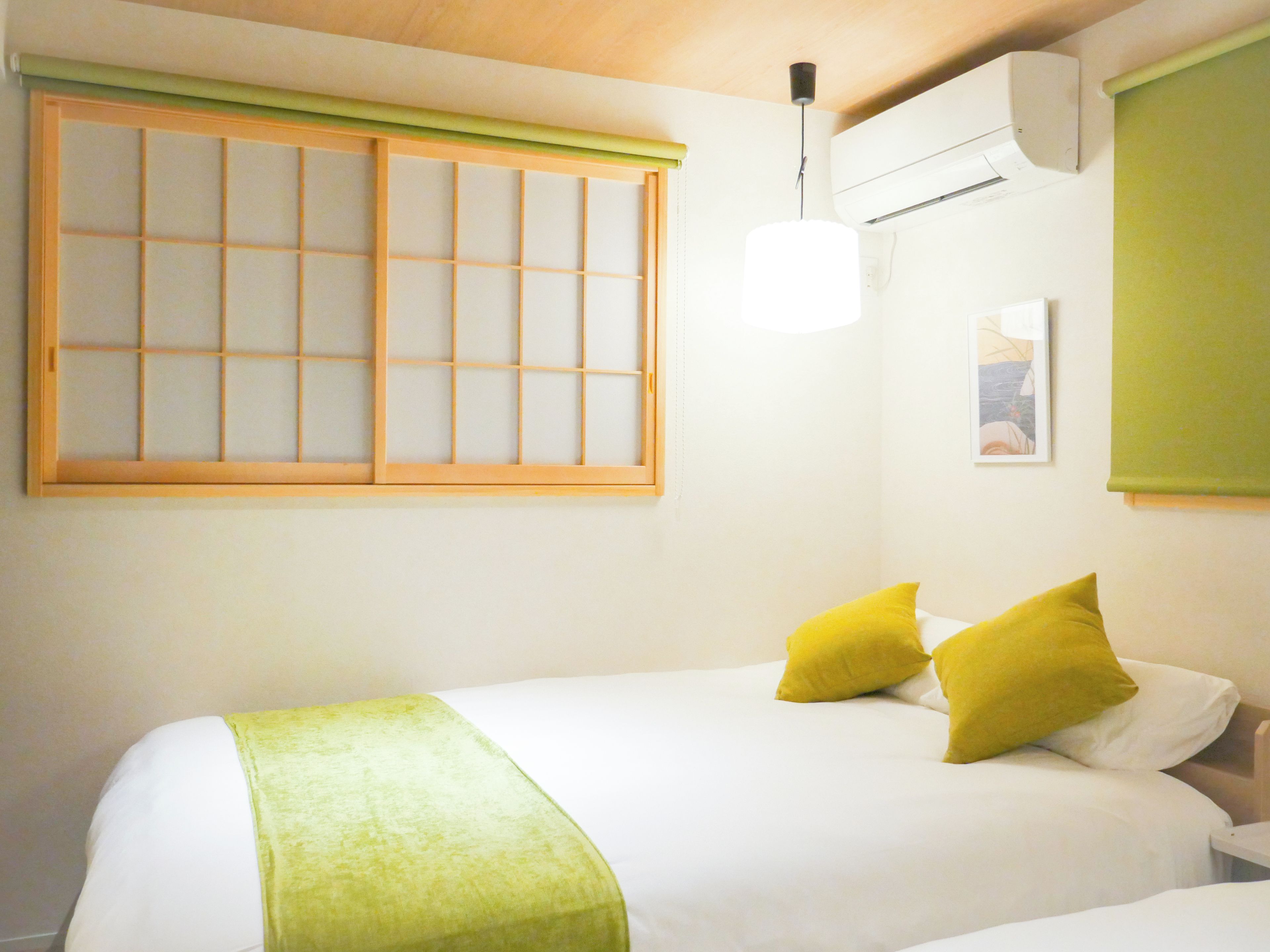 A hotel room with a bed, a window with a wooden frame, a wall-mounted air conditioner, and a bedside table.