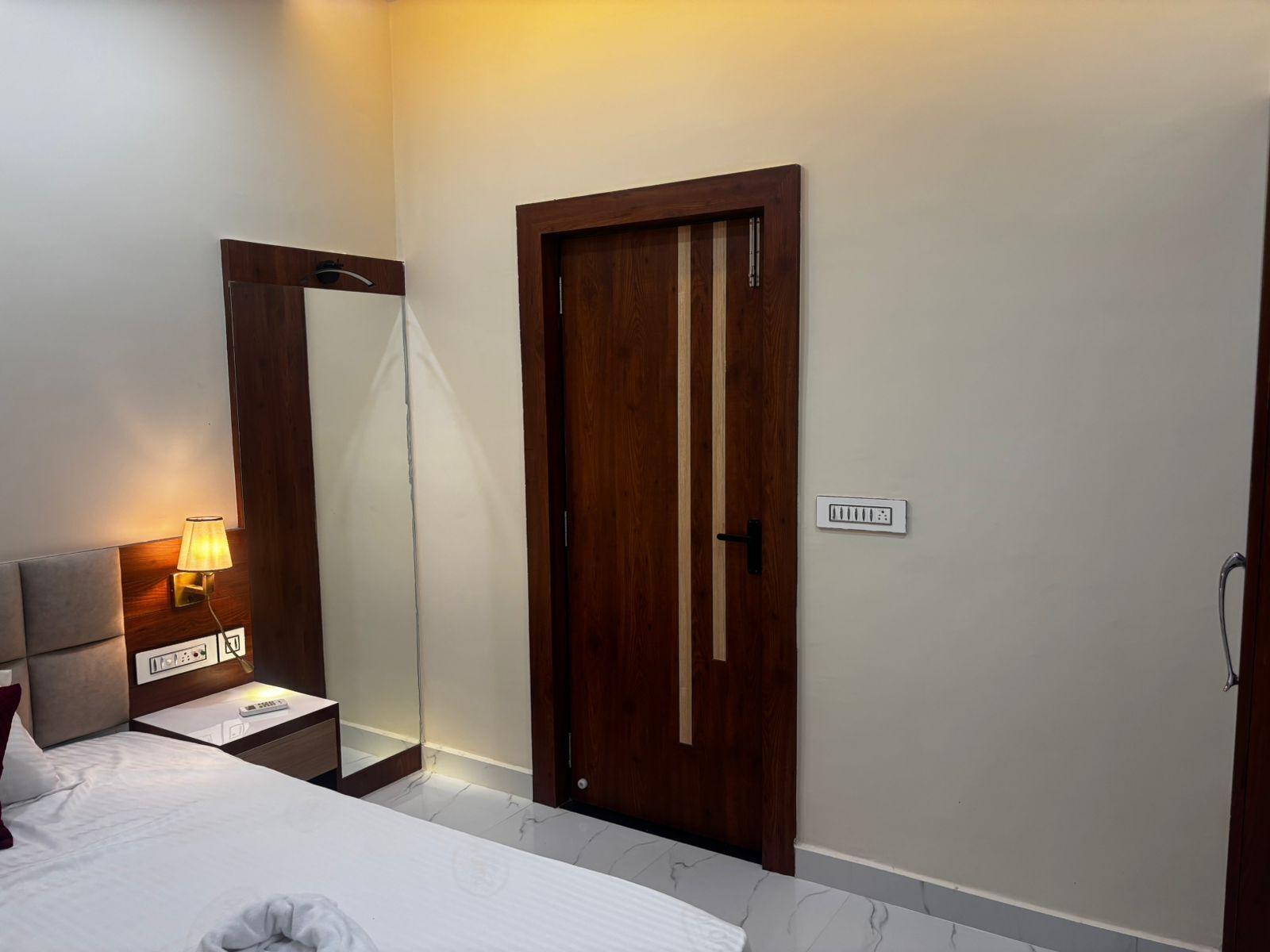 A hotel room with a bed, bedside table, lamp, mirror, wardrobe, and a power outlet.