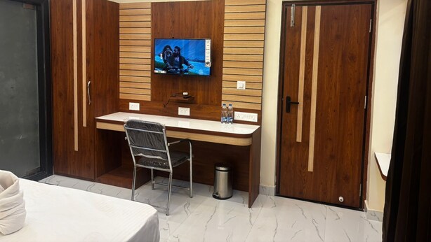 A hotel room with a wooden desk, a television mounted on the wall, a bed, and a wardrobe. - Asusss International (Kanthi)