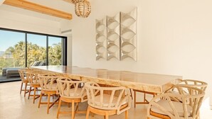 Un moderno comedor con una larga mesa de madera y sillas de mimbre, amplias ventanas con vistas a palmeras y una original obra de arte en la pared.