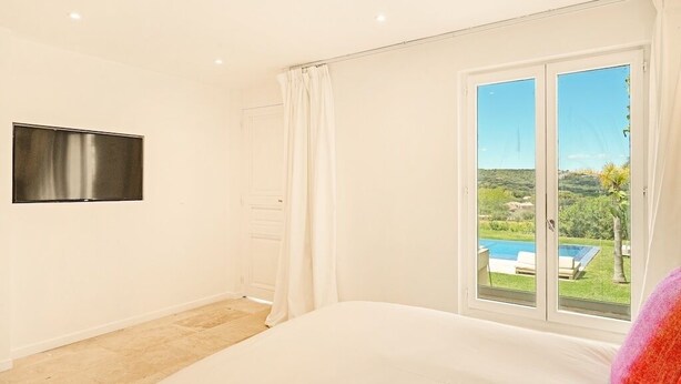 Un dormitorio con cama, televisor montado en la pared y puerta corrediza de vidrio que da a un balcón con vistas a una piscina y vegetación.