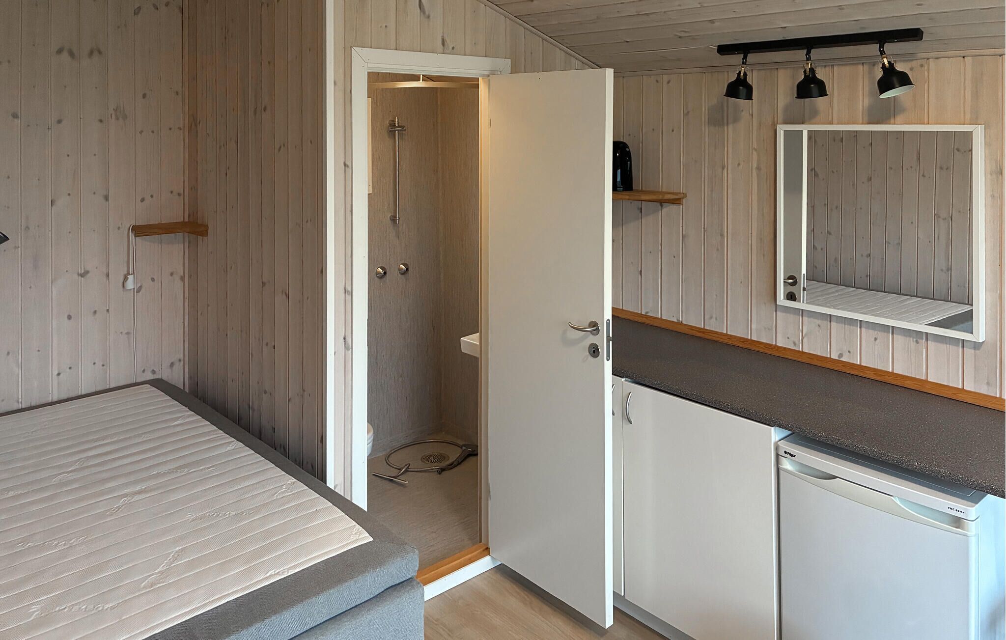 A compact room with a bed, a shower, a sink, and a mirror.