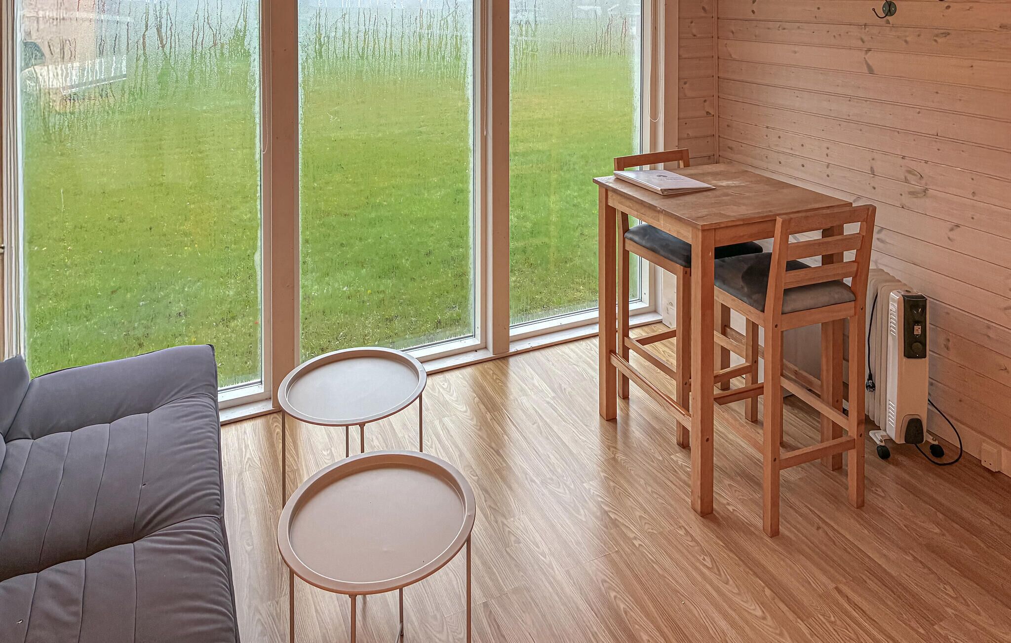 Een kamer met een houten tafel en stoelen, een kleine ronde tafel, een bank en uitzicht op een groen gazon door grote ramen.